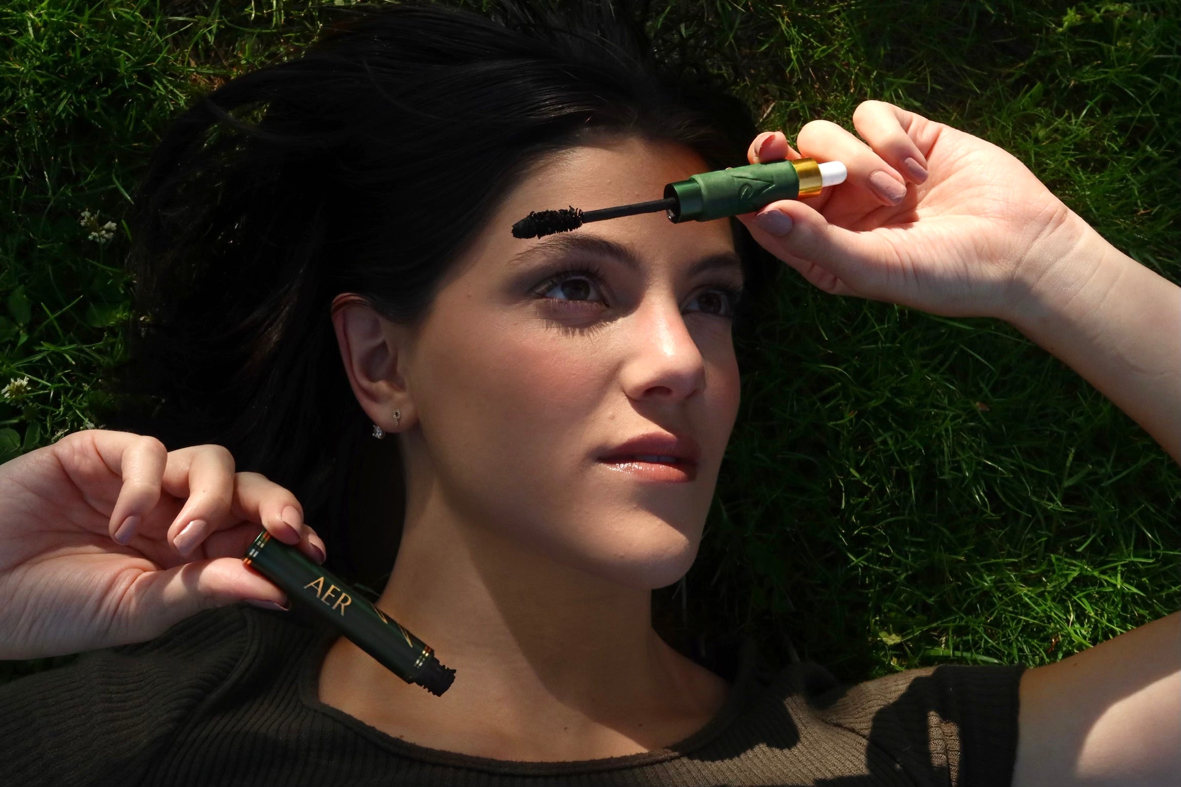 Paige laying in the grass holding her Aer Cosmetics mascara container. Photo credit to Honor Goldberg